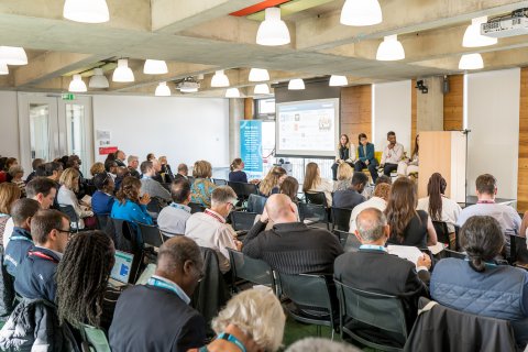 A photograph of the Annual Meeting taken from the back of the room with many people facing a stage with speakers on.